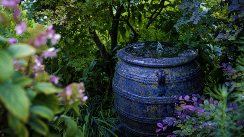 A dark blue bubbling urn fountain tucked in between trees and flowers in a garden.