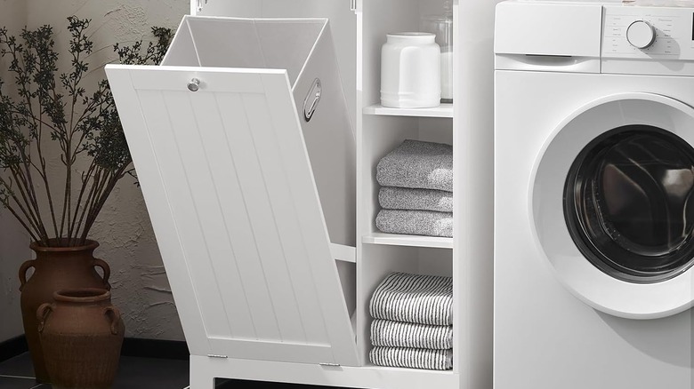 A tilt-out white laundry cabinet with shelves holding towels next to a washing machine. There is a plant in a brass pot to the left of the cabinet.
