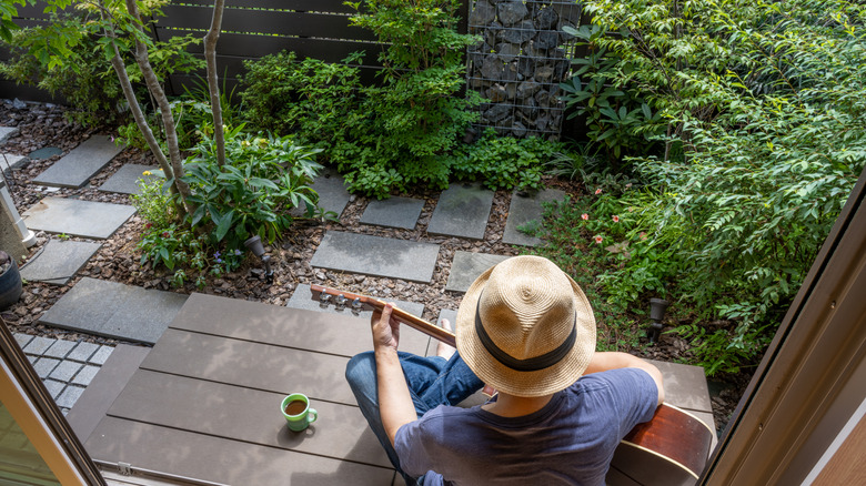 A person plays a guitar while sitting in their small backyard.