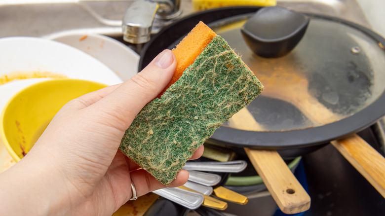 A person holds a dirty, old sponge in front of a pile of dirty dishes