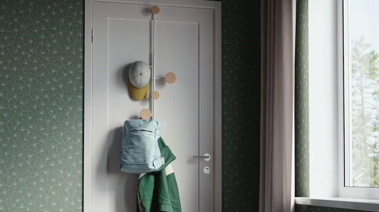 A BÄRFIS hanger hangs from a door in a bedroom and holds a hat, backpack, and jacket hanging from the knobs.