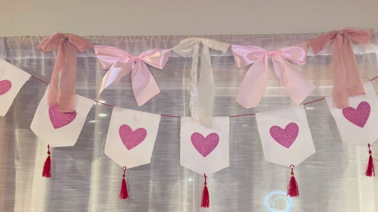 A screenshot of a video showing a garland made out of various bows hanging from a window curtain about a pre-made Valentine's Day garland