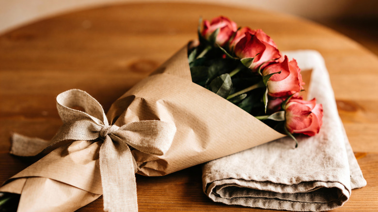 A paper-wrapped bouquet of roses with a burlap bow holding it together on a towel on top of a wooden surface