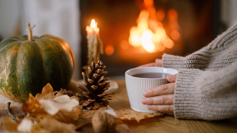 A woman wearing a sweater hiding a mug with pumpkins, a fire, acorns, and a candle in the background