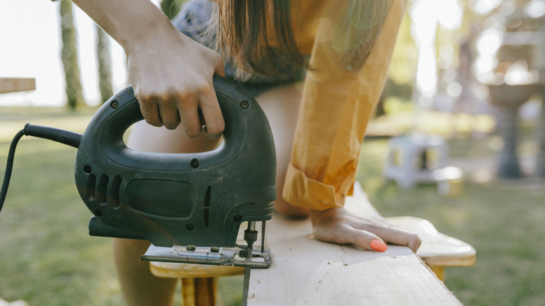 A woman with bright peach nails cutting through a piece of wood with a jigsaw tool