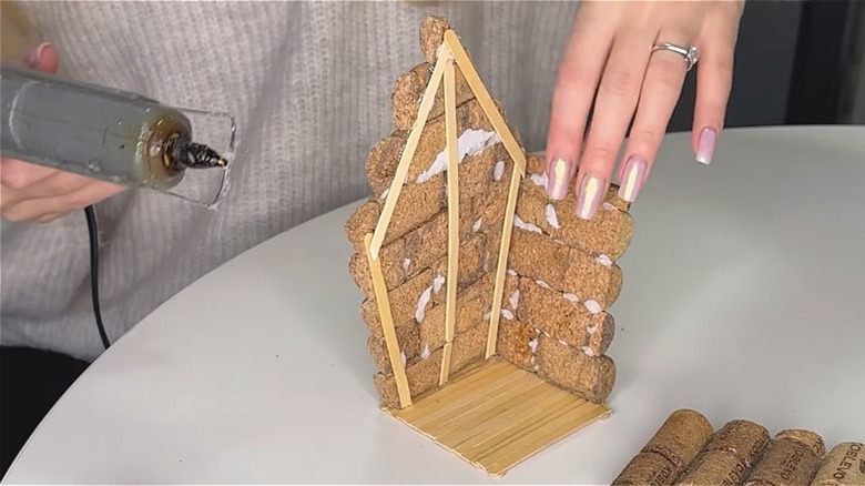 Person crafting a small bird house using corks, hot glue, and popsicle sticks.