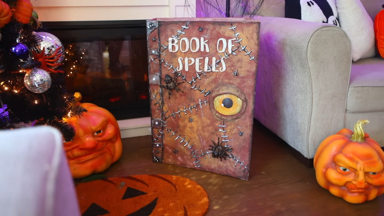 A large 'Book of Spells' Hallowen decor item rests by a fireplace with other seasonal decor.