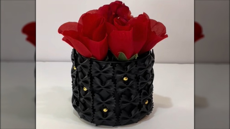 A glass vase decorated with farfalle pasta, gold accents, and black paint holds roses.