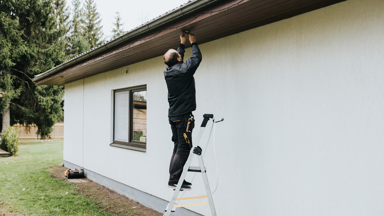 A light-skinned balding man with dark hair stands on a ladder to hang a hook from a roof overhang.