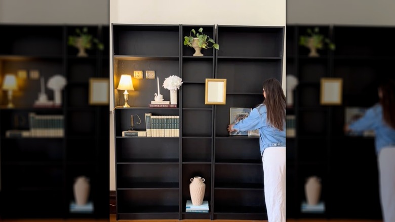 A woman arranges knickknacks on two large black bookshelves lining a wall in her home.