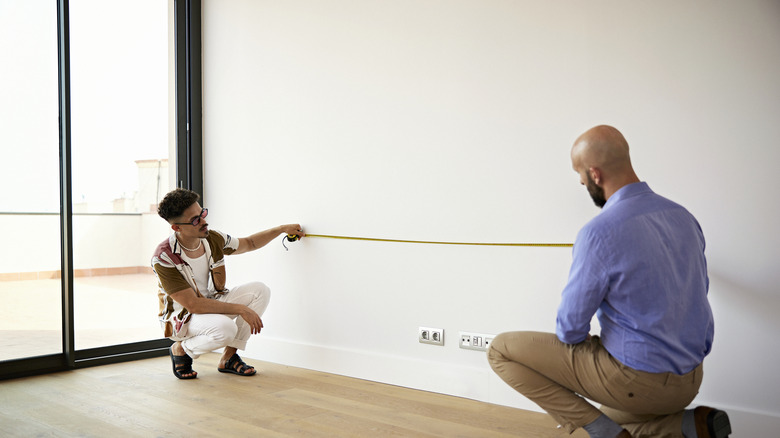 Two people measure the width of a bare white wall in an empty room in a condo.