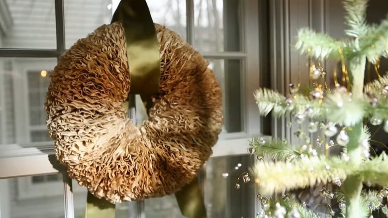 A wreath made from coffee filters hangs on a window from a wide gold ribbon looped through its middle.