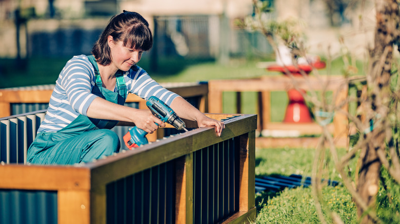 A woman uses a drill to build a raised garden bed out of metal and wood.