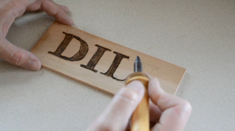 A wooden sign with "DILL" written in wood burning and person holding a pyrography tool