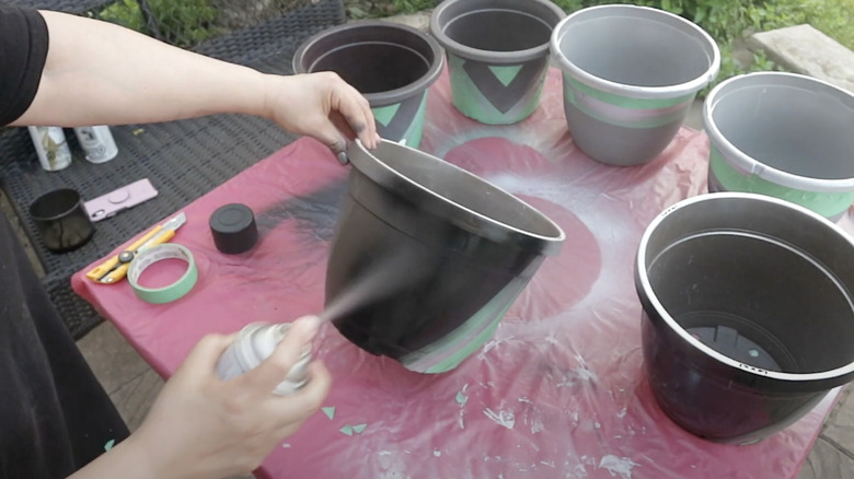 A person spray paints a plastic flower pot black outside on a table.