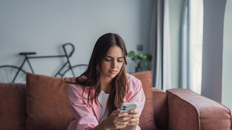 A woman sitting on a brown sofa and looking at her phone with a confused expression.
