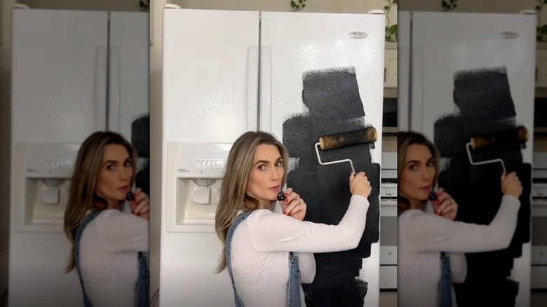 A woman using a paint roller to paint a white fridge black.