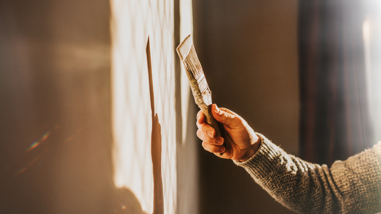 Sunlight shines through a window, creating a sun beam on an interior wall. A man holds a paintbrush and paints the wall a neutral color.