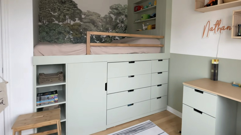 A lofted kid's bed with storage drawers and shelves underneath it and a tree mural.