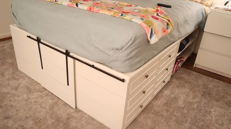 A bed with built-in storage has a mattress sitting atop a dresser next to shelving and barn door storage at the foot of the bed.