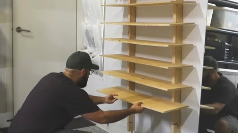 A man in a black baseball cap installing plywood vertical shelves into the white wall of a garage
