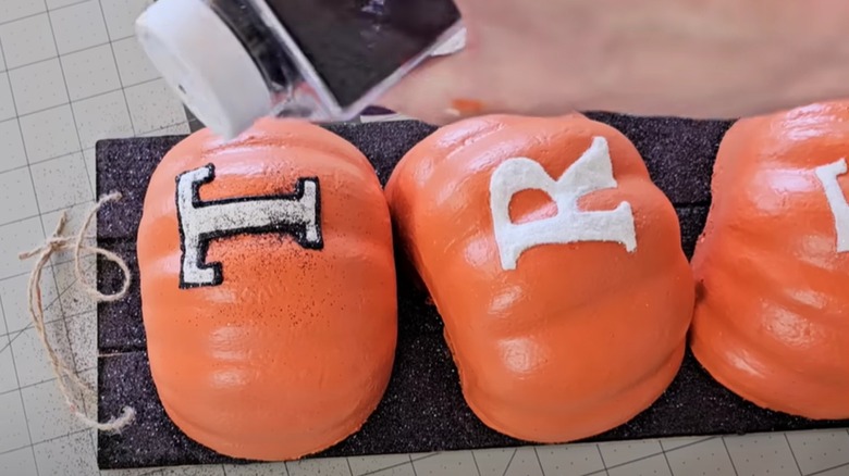 A hand sprinkles glitter on letters freshly painted on faux pumpkins.