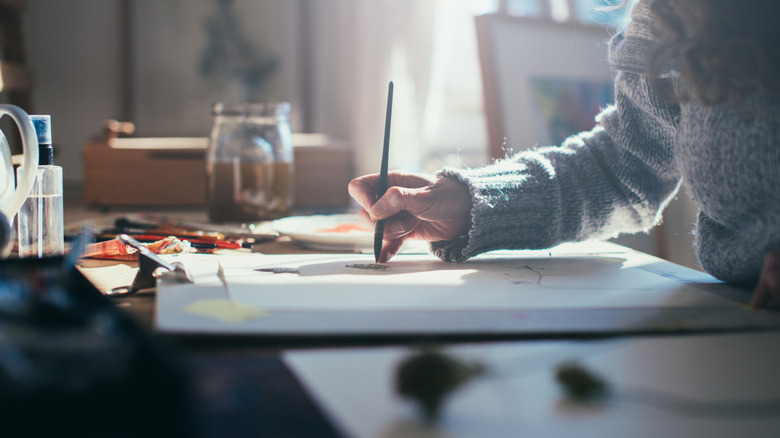 A woman in a grey sweater painting on a canvas with sunlight coming through the window.