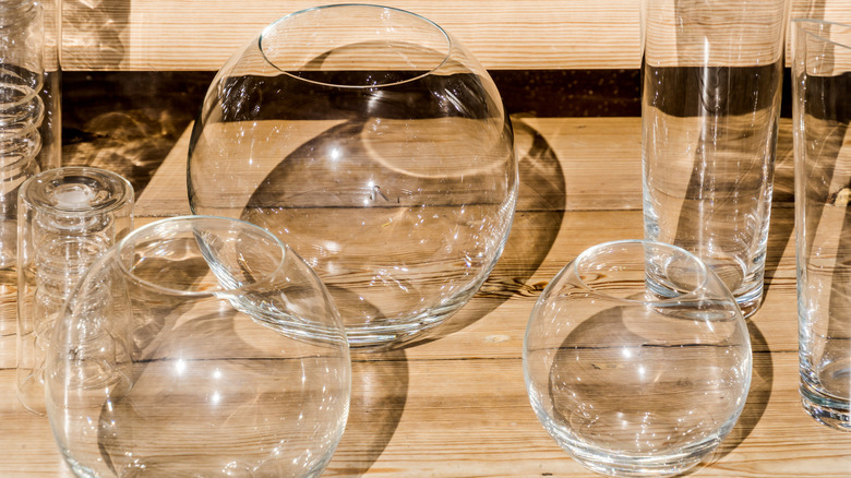 Clear round glass vases sit on a wooden shelf surrounded by other clear glassware