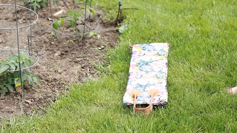 Floral fabric covered DIY kneeling pad laying in the grass near a vegetable garden