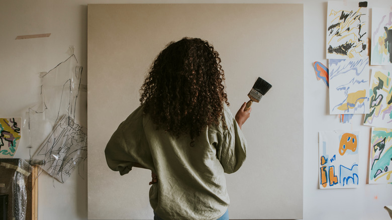 A woman holding a paintbrush stands in front of a blank canvas