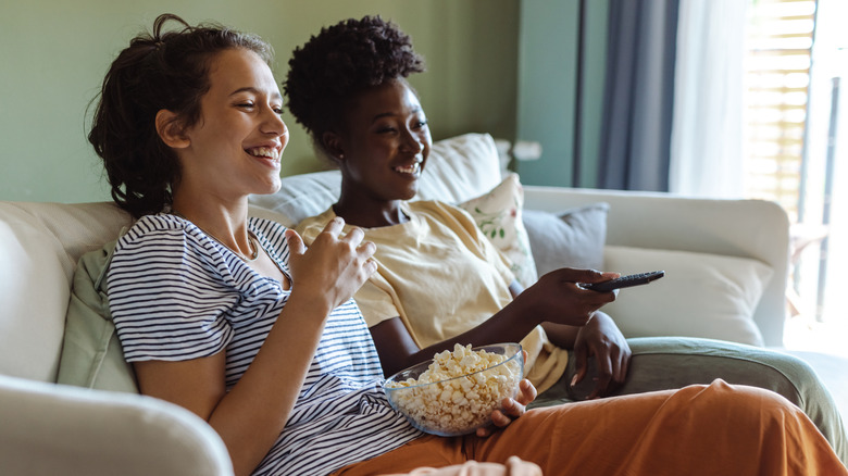 Two friends sitting on a couch at home watching a movie and eating popcorn.
