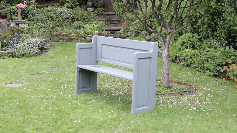 A gray wooden garden bench on a grassy area by some bushes and small tree