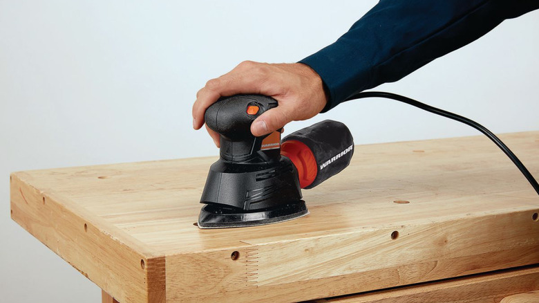 A hand covered in a dark-blue long sleeve using a Warrior palm sander to smoothen the top surface of a large slab of wood.