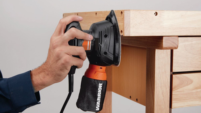 A hand holding the Warrior palm sander vertically while smoothing the edge of a wooden project