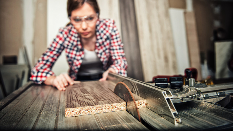 A person wearing protective goggles runs a wood plank through a table saw.