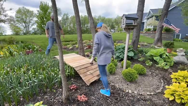 A couple position a DIY wood bridge in a landscaped garden.