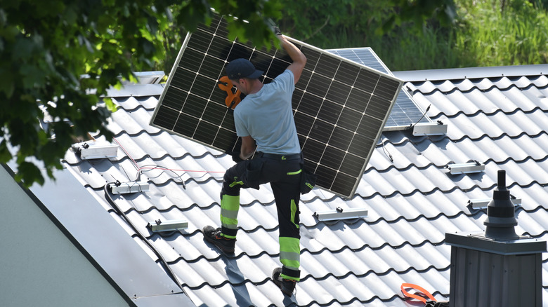 A person standing on a roof, holding a solar panel, in the process of installing it