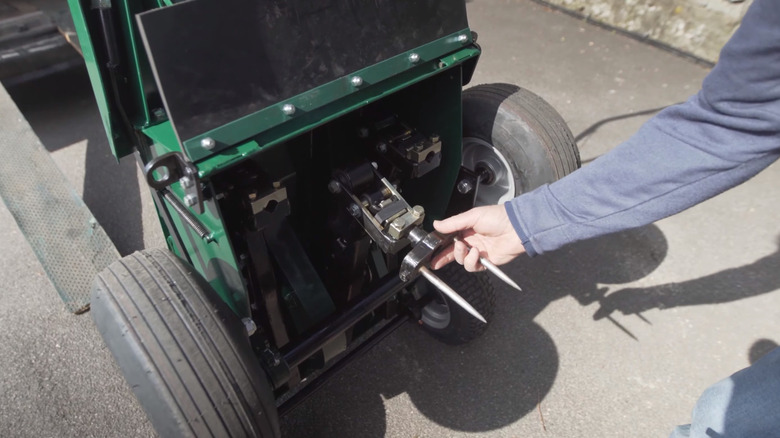A hand reaching to grab two aerator tines from the bottom of a machine