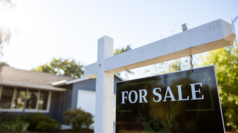 Close-up of a black and white 'for sale' sign with a blurred house in the background