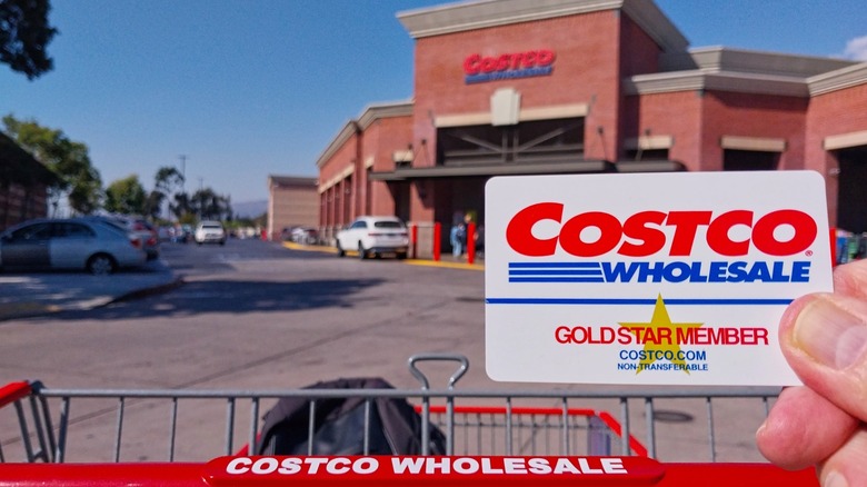 A Costco Wholesale Gold Star Member card held up in front of a Costco in the distance.