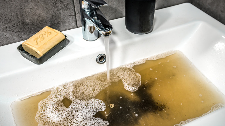 A clogged sink with a running tap and dirty brown water