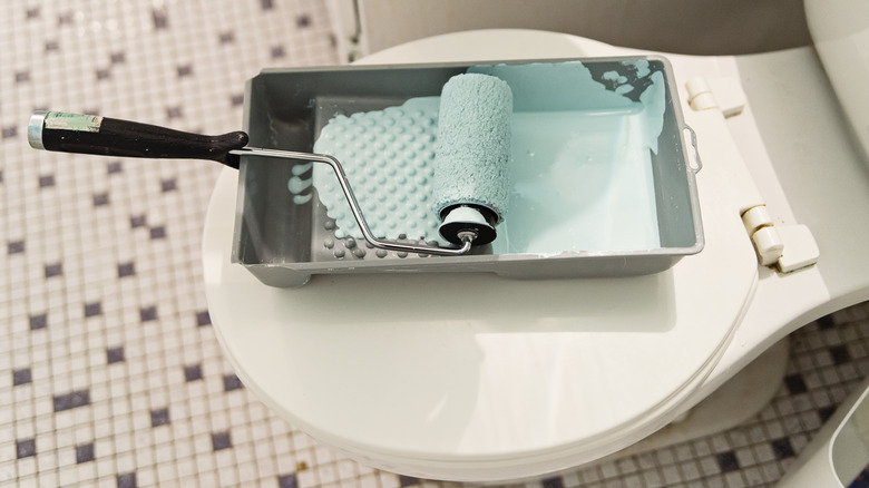 Light green paint in a tray with a roller sitting on a toilet lid with a tiled bathroom floor in the background.