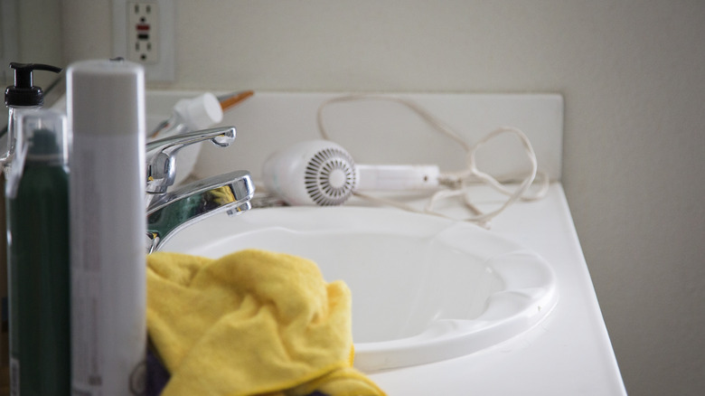 Cluttered bathroom countertop with a hairdryer, products, and a face towel