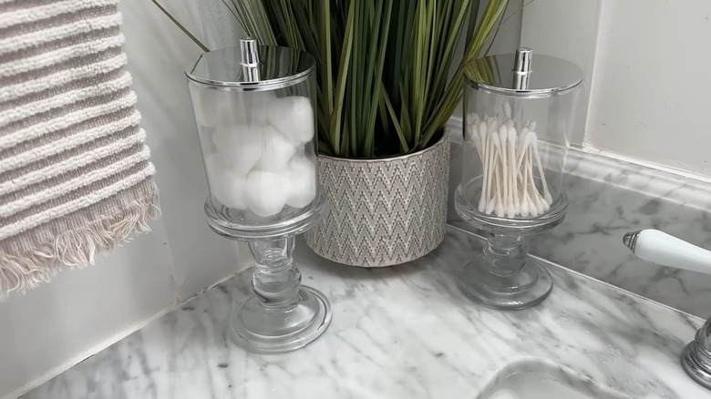 Two clear storage containers raised by candle holders with cotton balls and cotton swabs inside, sitting on a bathroom countertop