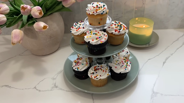 Two plates with candle holder pedestals create a cupcake stand sitting on a countertop next to a candle and flowers