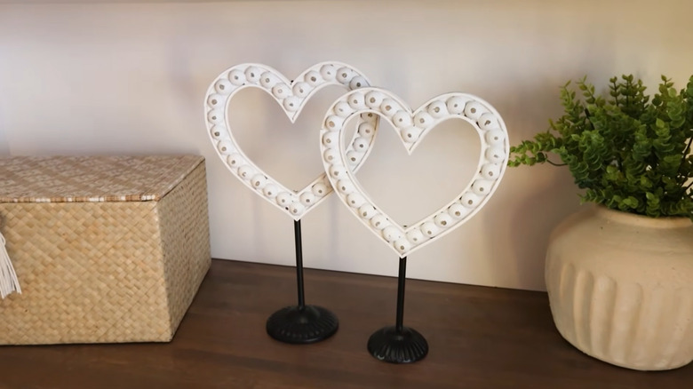 Two antiqued wooden heart cutouts attached to black metal candlestick holders sitting on a table near other decor