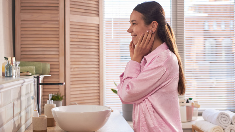 Woman in pink pajamas looks at herself in the bathroom mirror.