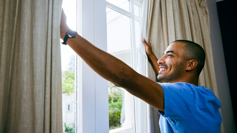 A smiling African American man parts heavy window curtains to look outside.