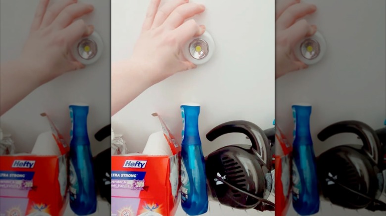 A person places a push light on a white wall above some cleaning supplies.