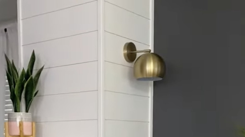 The wall sconce is attached to a white shiplap wall with a grey wall and plant on either side